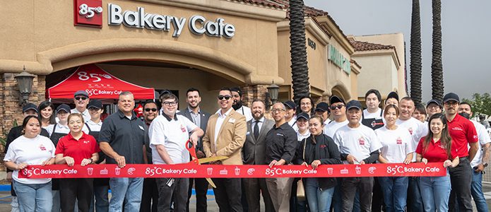 A group of people, including bakery employees and officials, stand together in front of 85°C Bakery Cafe, holding a red ribbon during a ribbon-cutting ceremony.