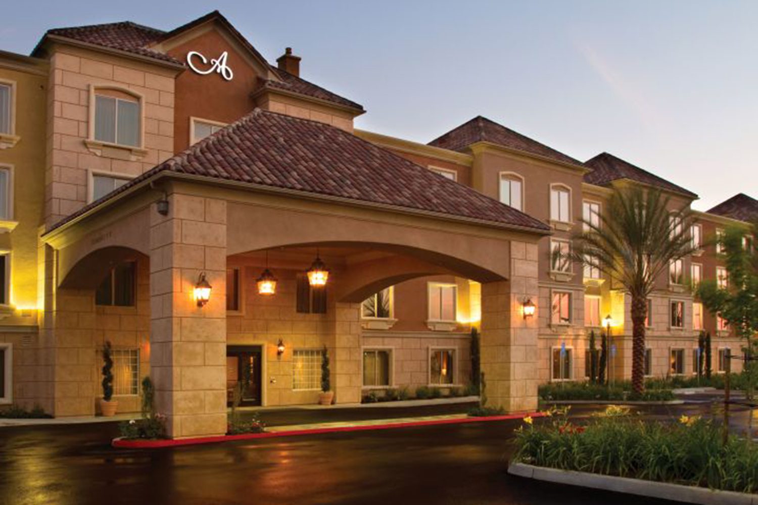 A beige, multi-story building with a tile roof and an illuminated entrance stands at dusk, featuring a portico and palm trees near the entrance.