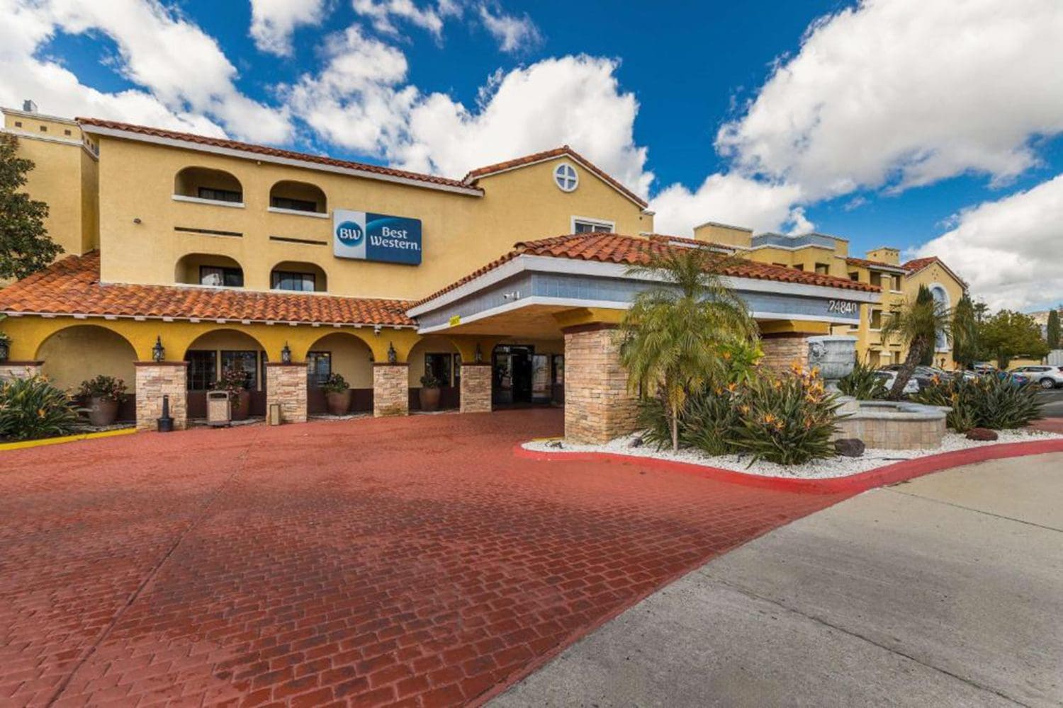 Front view of a Best Western hotel with a red brick driveway, stone pillars, palm trees, and a clear blue sky with scattered clouds.