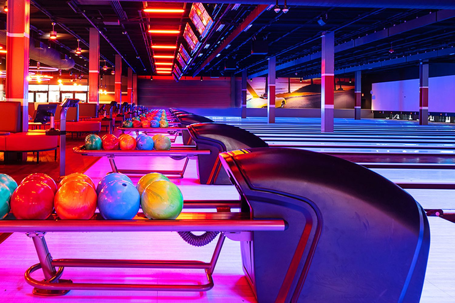 A brightly lit bowling alley with multiple lanes, colorful bowling balls arranged in racks, and electronic scoring screens overhead.