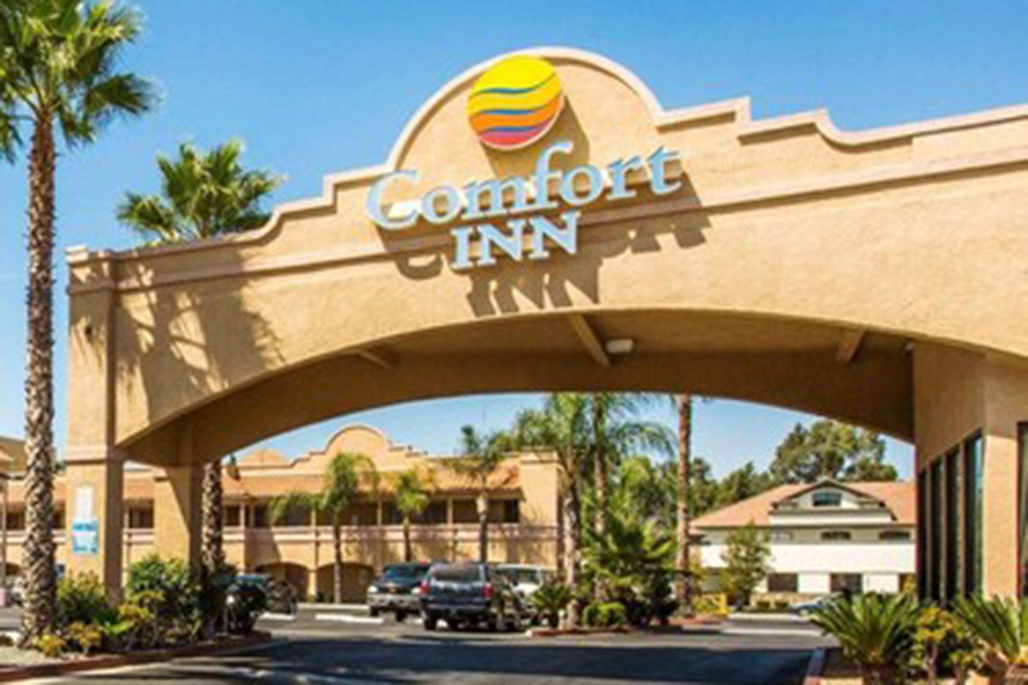 The image shows the entrance of a Comfort Inn hotel with an archway displaying the hotel's name and logo. Palm trees and parked cars are visible in the parking lot.