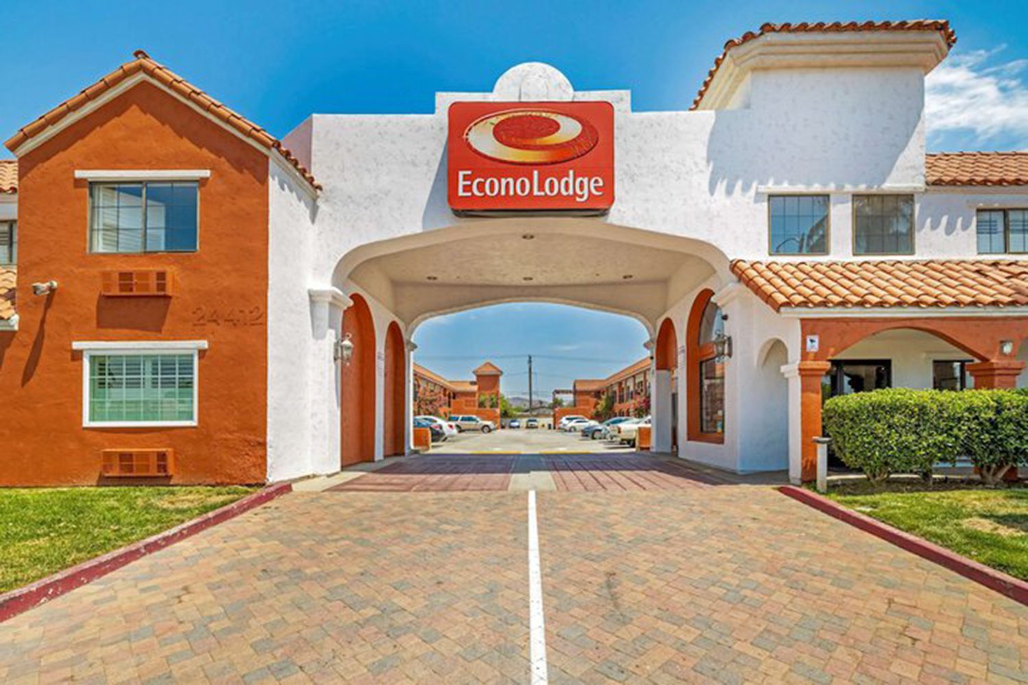 The image shows the entrance of an Econo Lodge hotel with a white archway and orange roof tiles. There is a sign with the hotel logo above the arch and a paved driveway leading to the parking area.