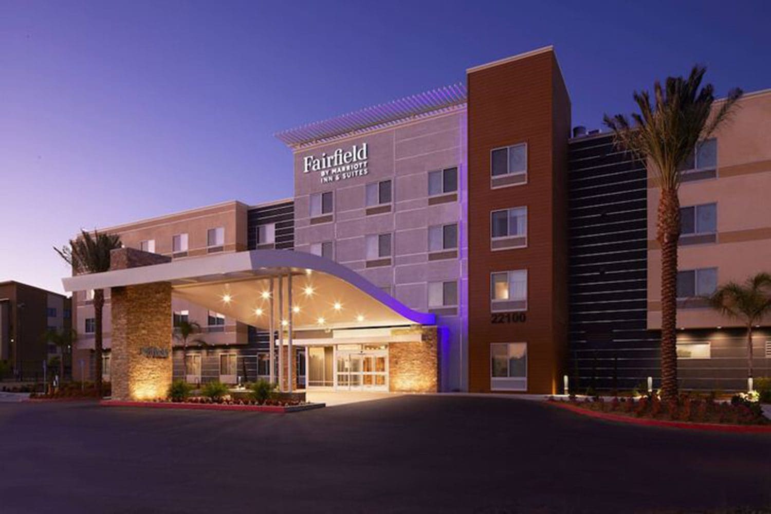 A modern hotel exterior with a covered entrance, palm trees, and well-lit signage reading "Fairfield Inn & Suites" at dusk.