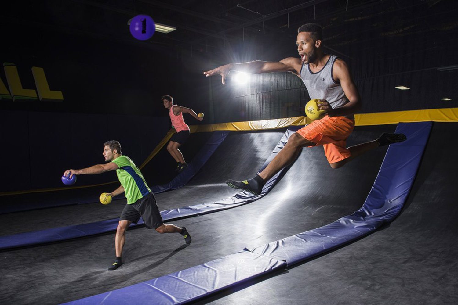 Three people in athletic clothing play dodgeball on an indoor trampoline court, mid-jump and holding dodgeballs, lit by overhead lighting.