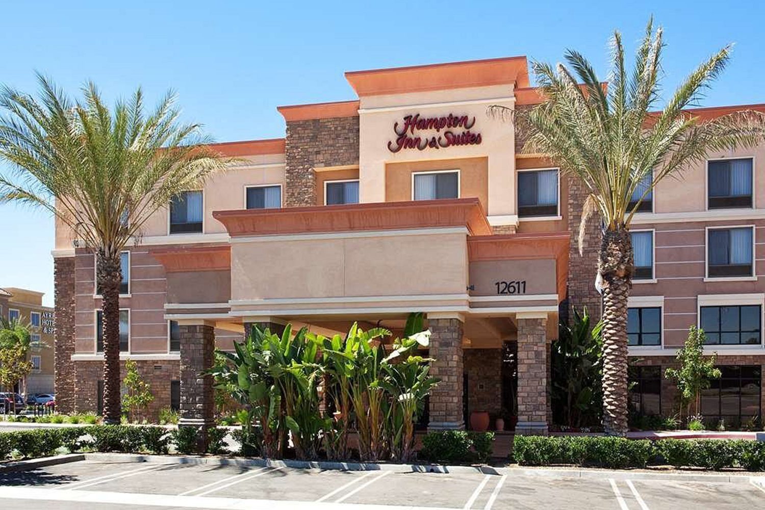 Exterior view of a Hampton Inn & Suites hotel with surrounding palm trees and a clear blue sky in the background.