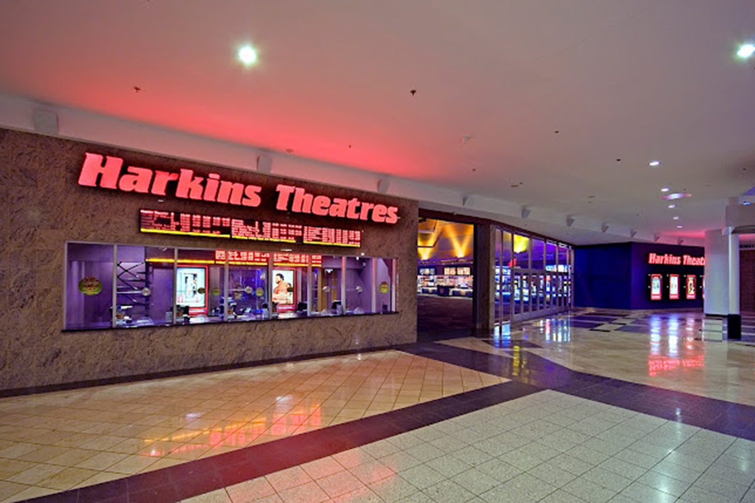 Image showing the entrance to Harkins Theatres inside a shopping mall, with box office counters illuminated by pink-red neon signage. The mall is spacious with a polished tile floor.