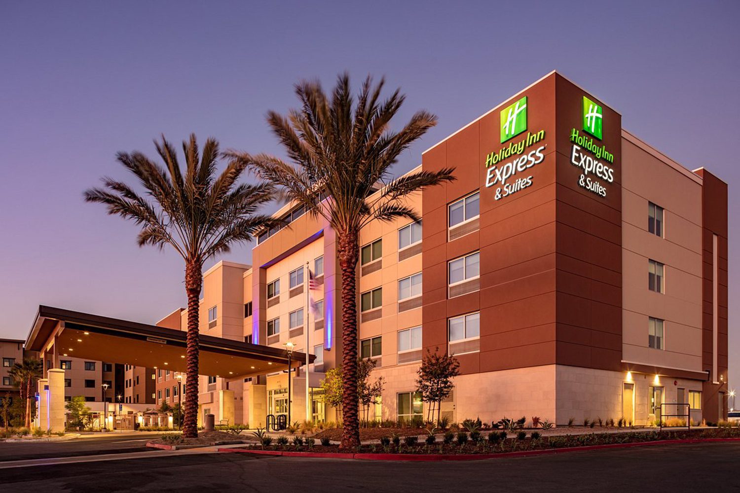 Exterior view of a Holiday Inn Express & Suites hotel at dusk, with illuminated signs and palm trees flanking the entrance.