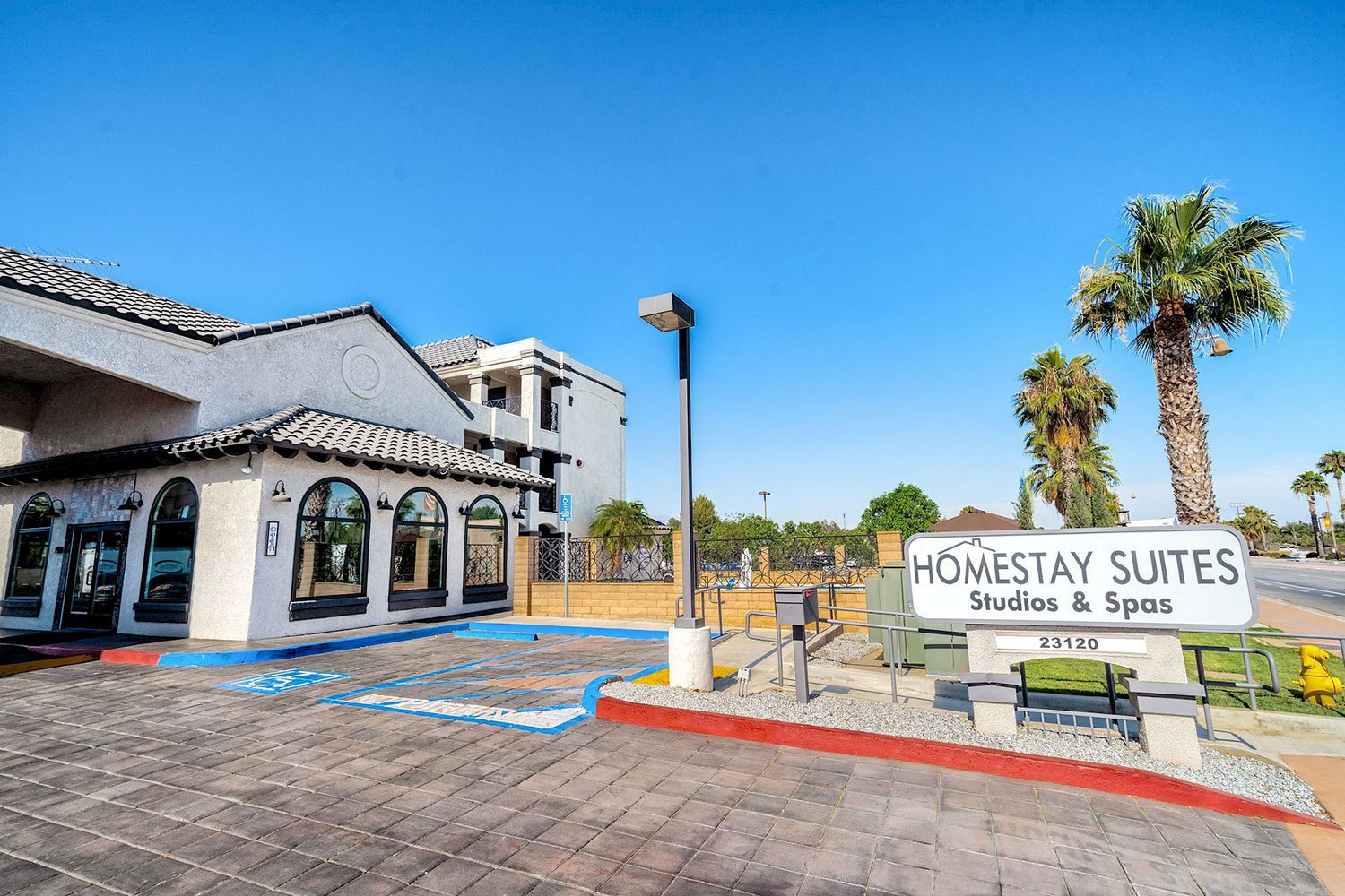 A hotel named "Homestay Suites Studios & Spas" with a clear entrance, parking area, and palm trees in the background under a clear blue sky.