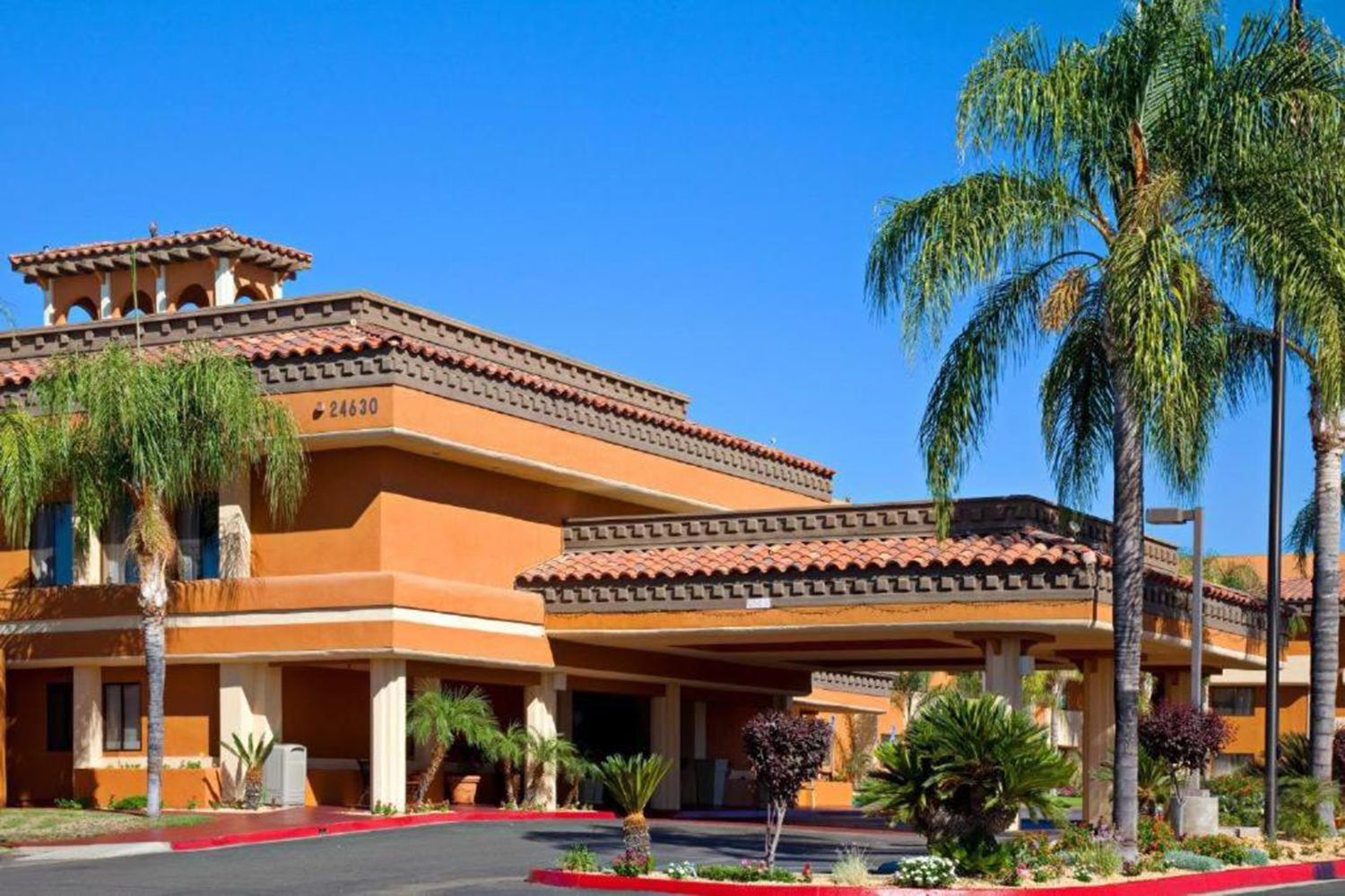 A two-story building with a terra-cotta roof, palm trees, and well-maintained landscaping, under a clear blue sky. The facade is painted in shades of ochre and brown.