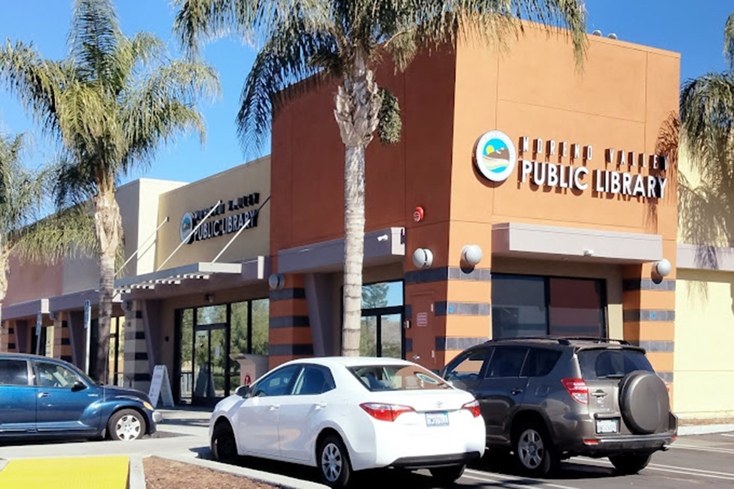 A public library building with a sign labeled "Public Library" in front. Several cars are parked in the lot outside, and palm trees are visible.