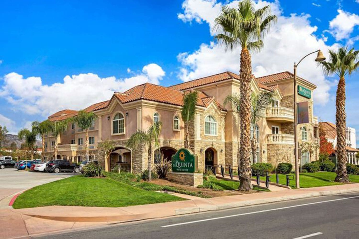 A tan-colored La Quinta Inn & Suites hotel with a tile roof, palm trees, and several parked cars under a bright blue sky with white clouds.