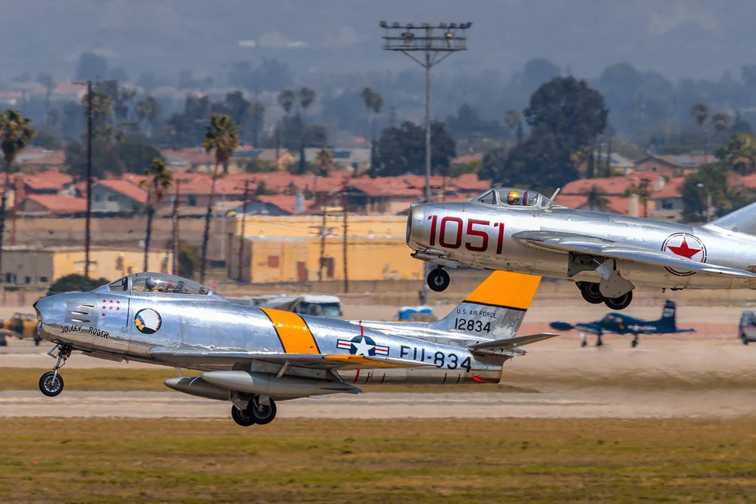 Two vintage fighter jets, one with the identifier "FU-834" and another with "1051," take off from a runway with buildings and palm trees in the background.