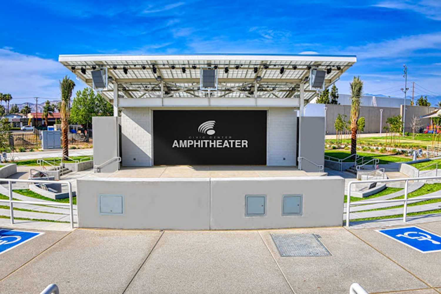 Outdoor amphitheater with a modern stage, overhead structure, and seating area. The word "Amphitheater" displayed prominently on the stage's backdrop. Accessible parking spaces in the foreground.