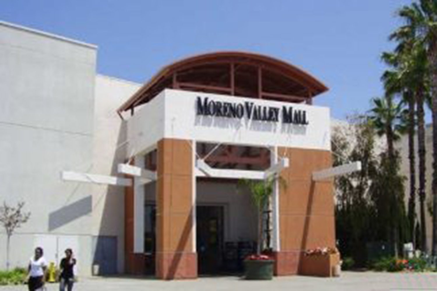 Entrance to Moreno Valley Mall with a clear, sunny sky and palm trees in the background. Two people are walking near the mall's entrance.