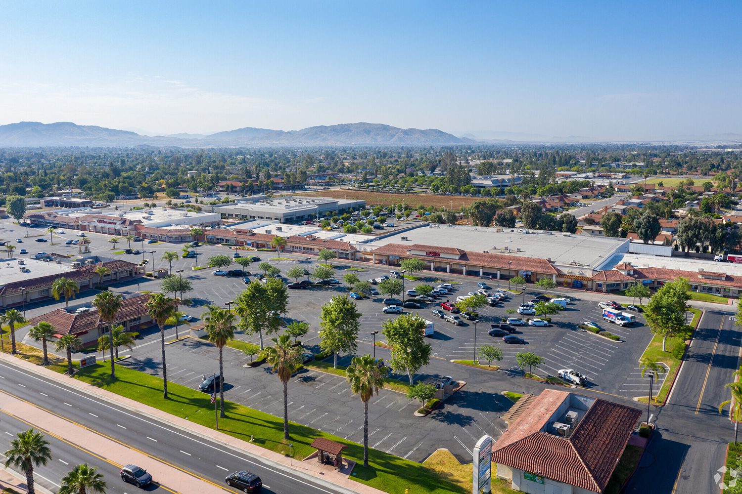 Aerial view of a large shopping center parking lot with scattered cars, surrounded by buildings, palm trees, and mountains in the background on a sunny day.