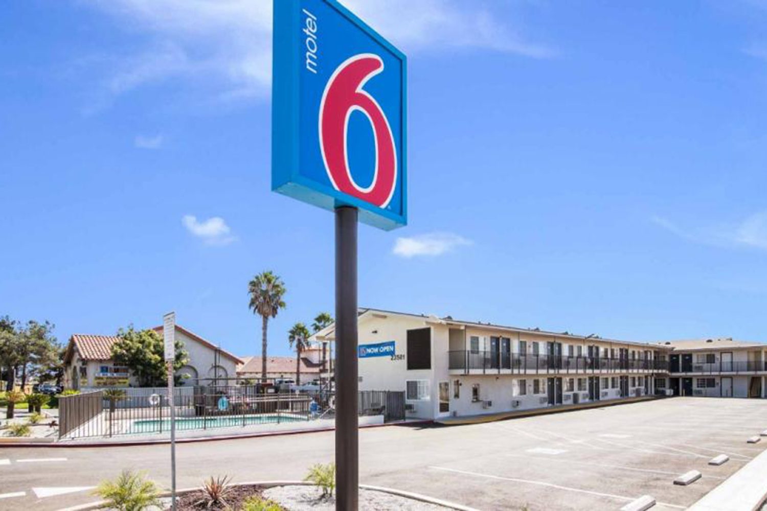 Exterior view of a Motel 6 building with a parking lot in front, a sign displaying the motel's name, and an outdoor swimming pool on the left under a clear blue sky.