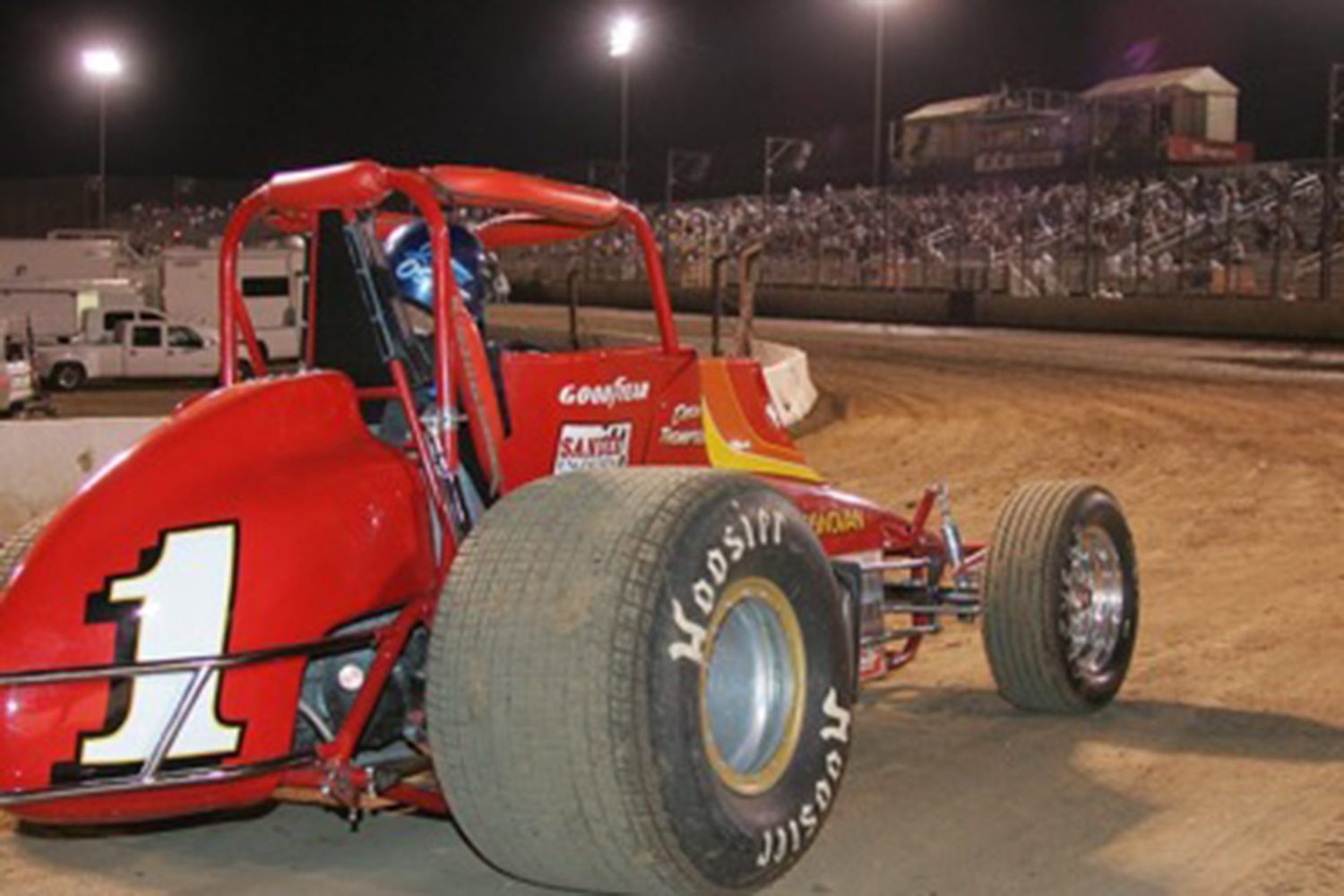 A red race car with the number 1 is on a dirt track at night, with the stands and other cars in the background.