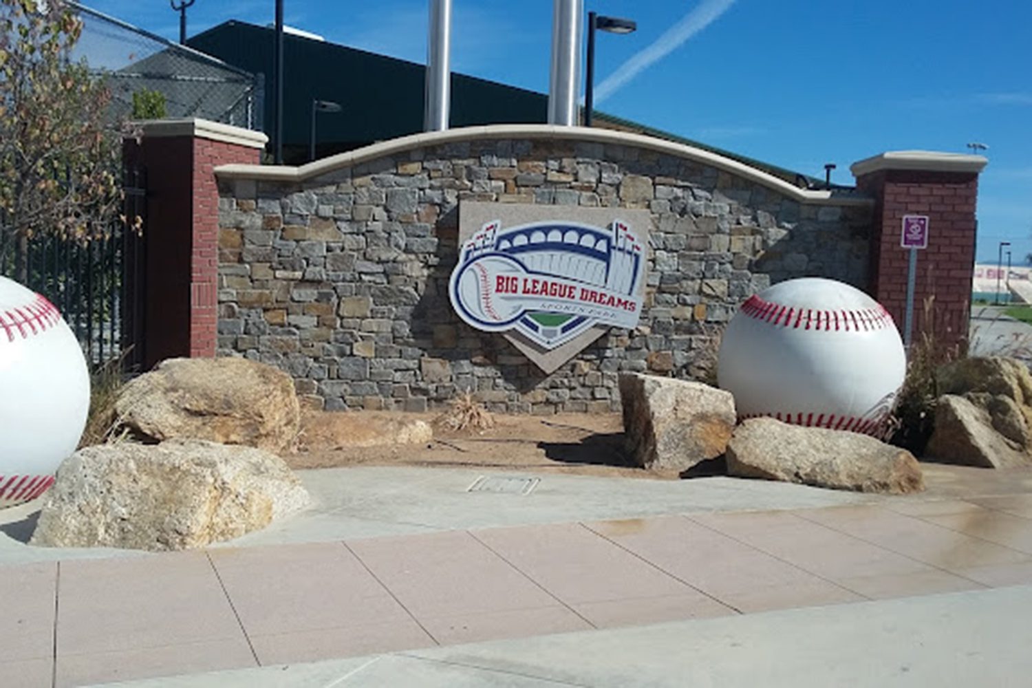 Entrance monument for Big League Dreams with a sign featuring a baseball stadium logo, flanked by two large baseball sculptures and surrounded by rocks, set against a stone wall background.