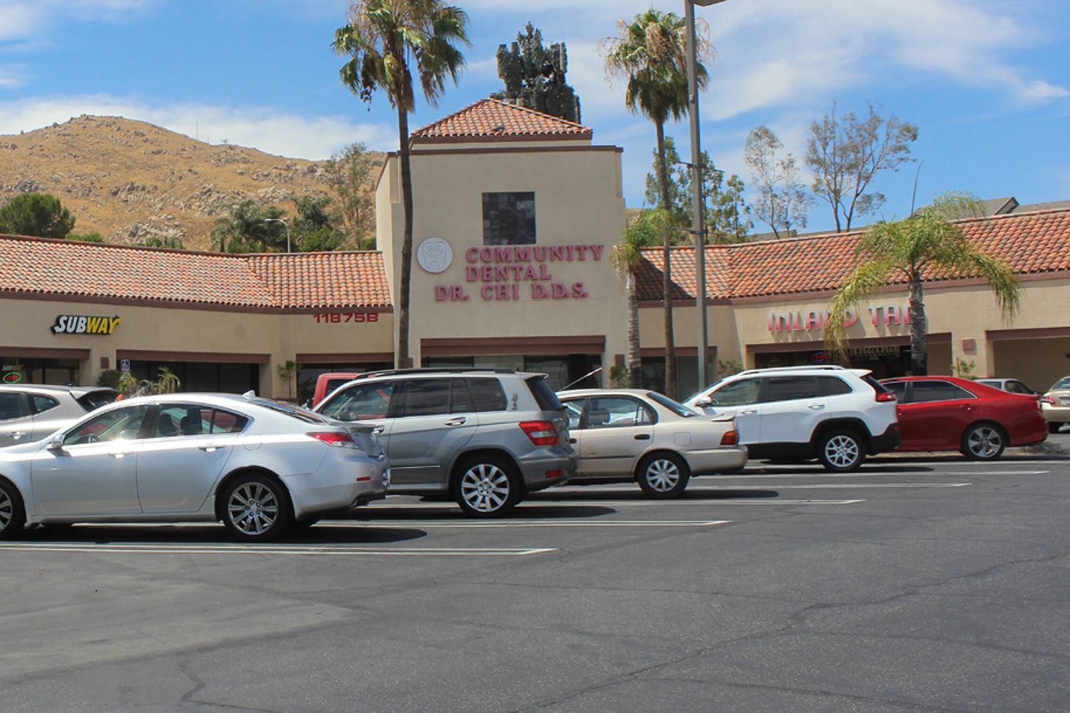 A strip mall parking lot with several parked cars in front of businesses, including a Community Dental office, a nail salon, and a Subway sandwich shop on a sunny day.