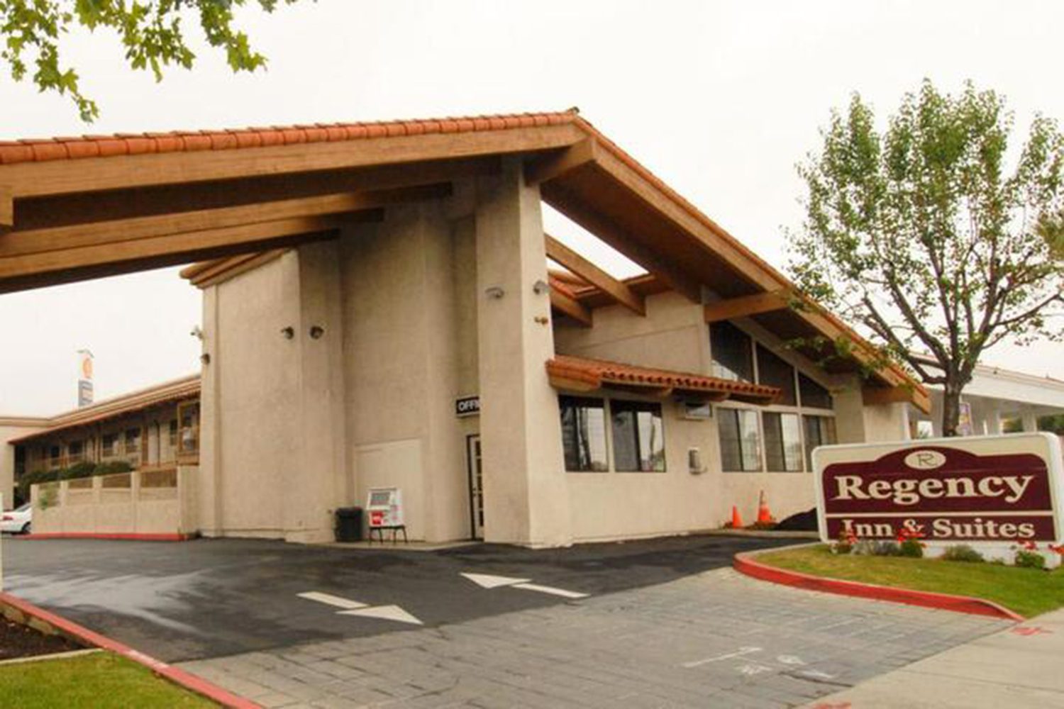 A beige building with a wooden roof structure houses the Regency Inn & Suites. A sign with the hotel name stands in the foreground near a tree. An entrance driveway is visible.