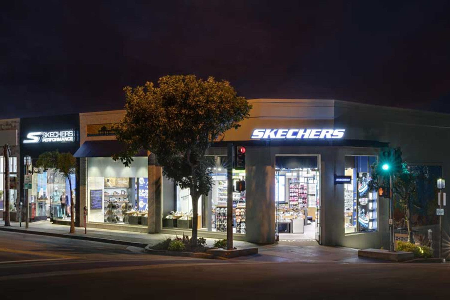 Night view of a Skechers retail store with illuminated signage and storefront displays, located at a street intersection with traffic lights and trees.