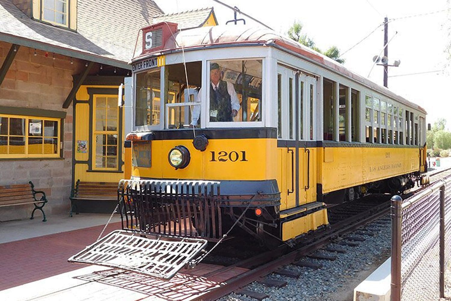 A yellow vintage streetcar numbered 1201 is parked next to a historical train station with a brick building and benches. A driver is visible in the front cabin.