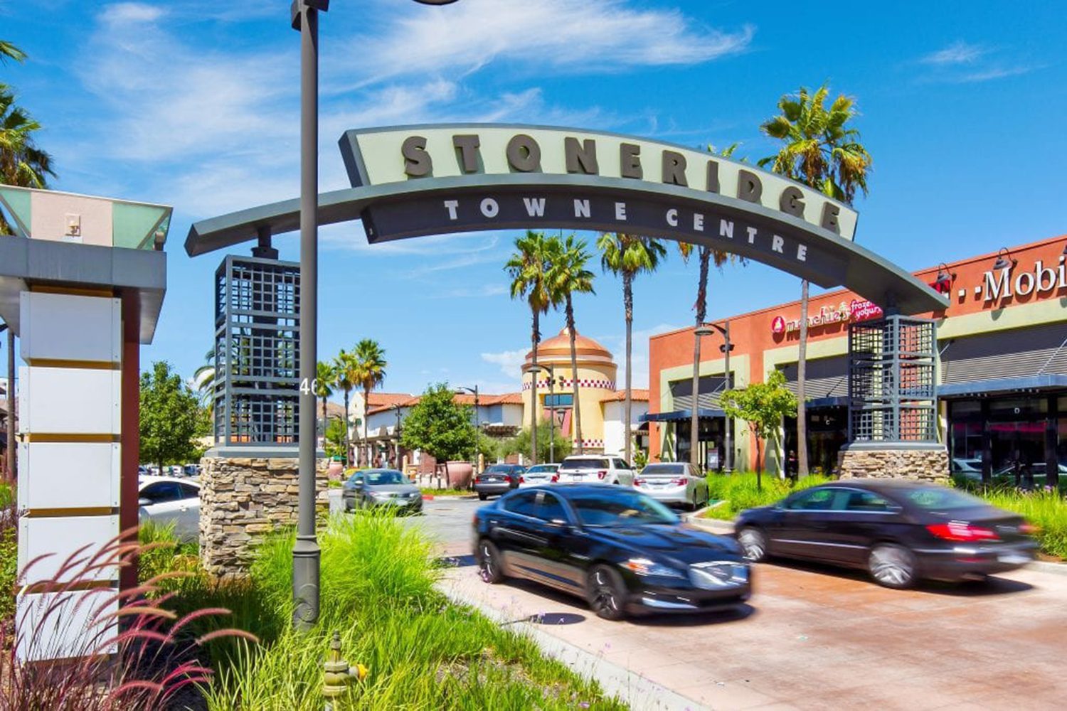 Entrance to Stoneridge Towne Centre with cars on the road, shops, and palm trees in the background under a clear sky.