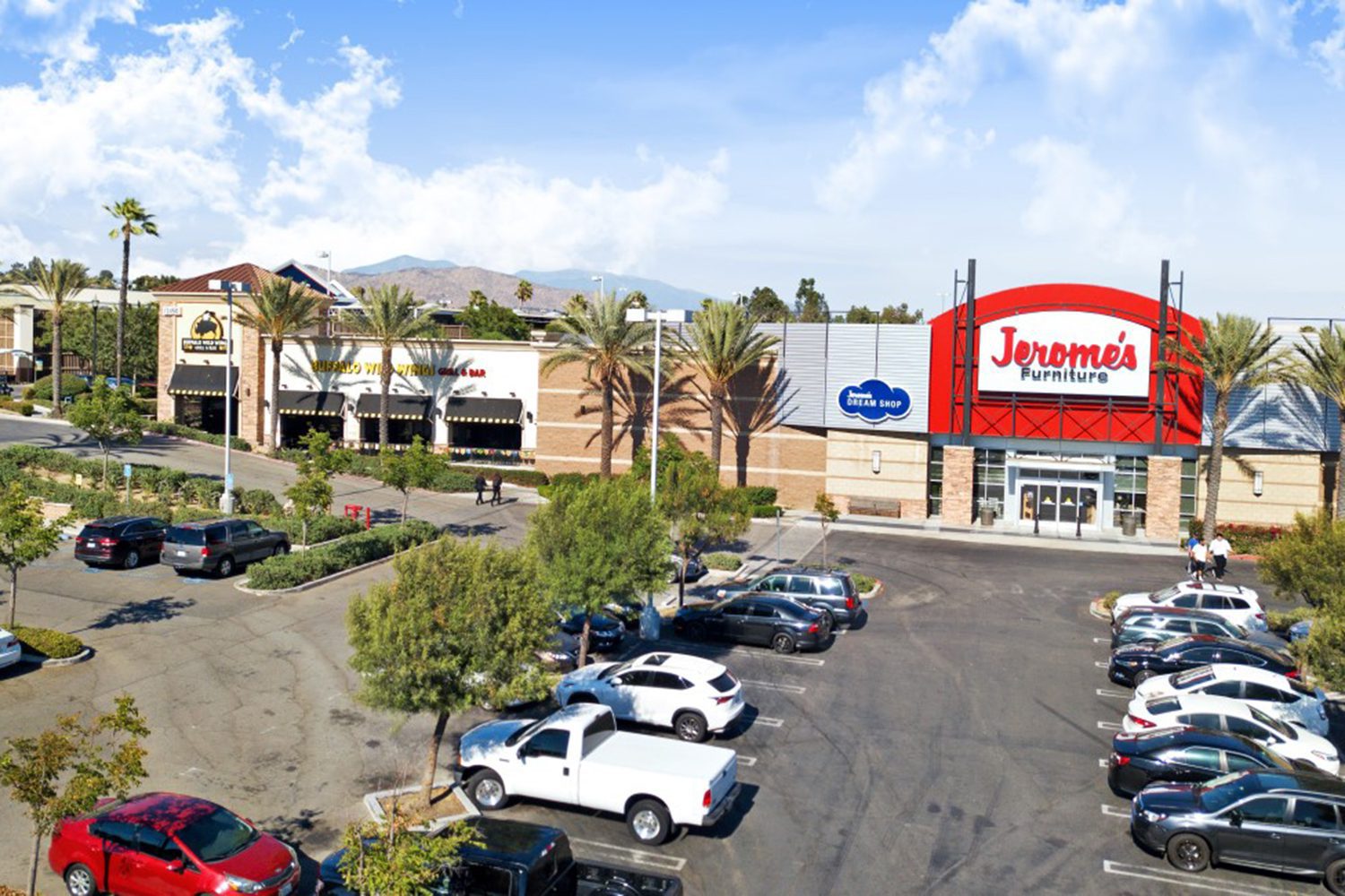 A parking lot outside a shopping center with a Jerome's Furniture store and several parked vehicles. Palm trees and additional shops are visible under a partly cloudy sky.