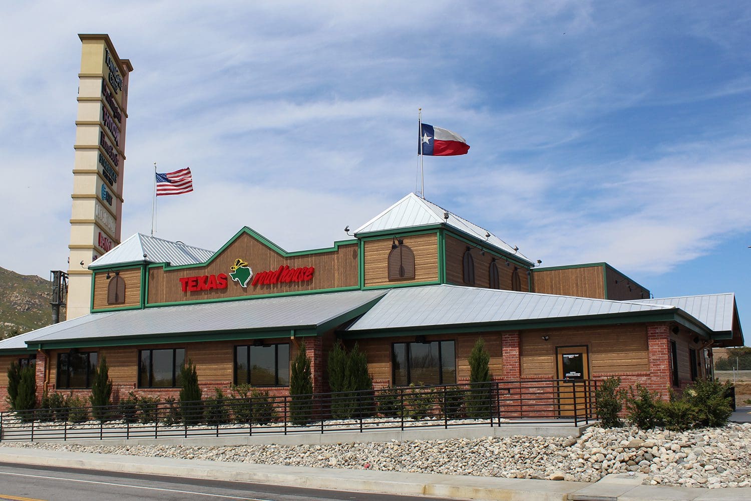 A Texas Roadhouse restaurant with an American and Texas state flag on display, featuring a rustic exterior with wooden paneling and a green roof, set against a backdrop of a clear blue sky.