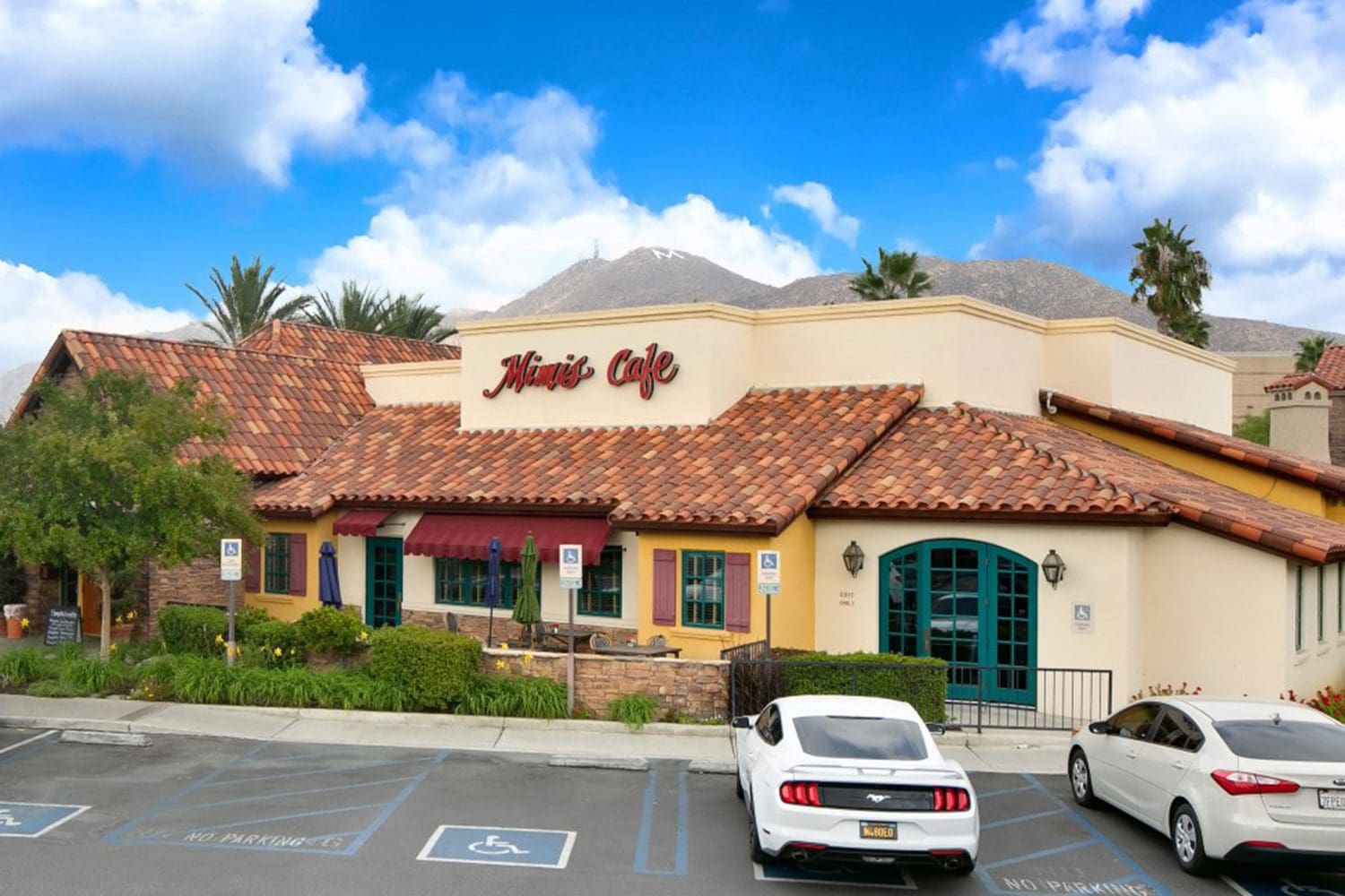 A beige stucco building with a red tiled roof houses "Mirage Cafe." Several cars are parked in front, and blue sky with white clouds is visible in the background.