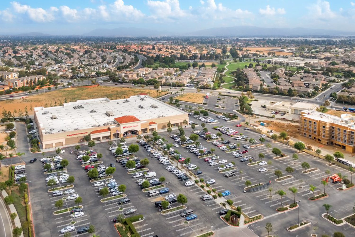 Aerial view of a shopping center with a large store and parking lot filled with cars, surrounded by residential area and landscape of houses and greenery.