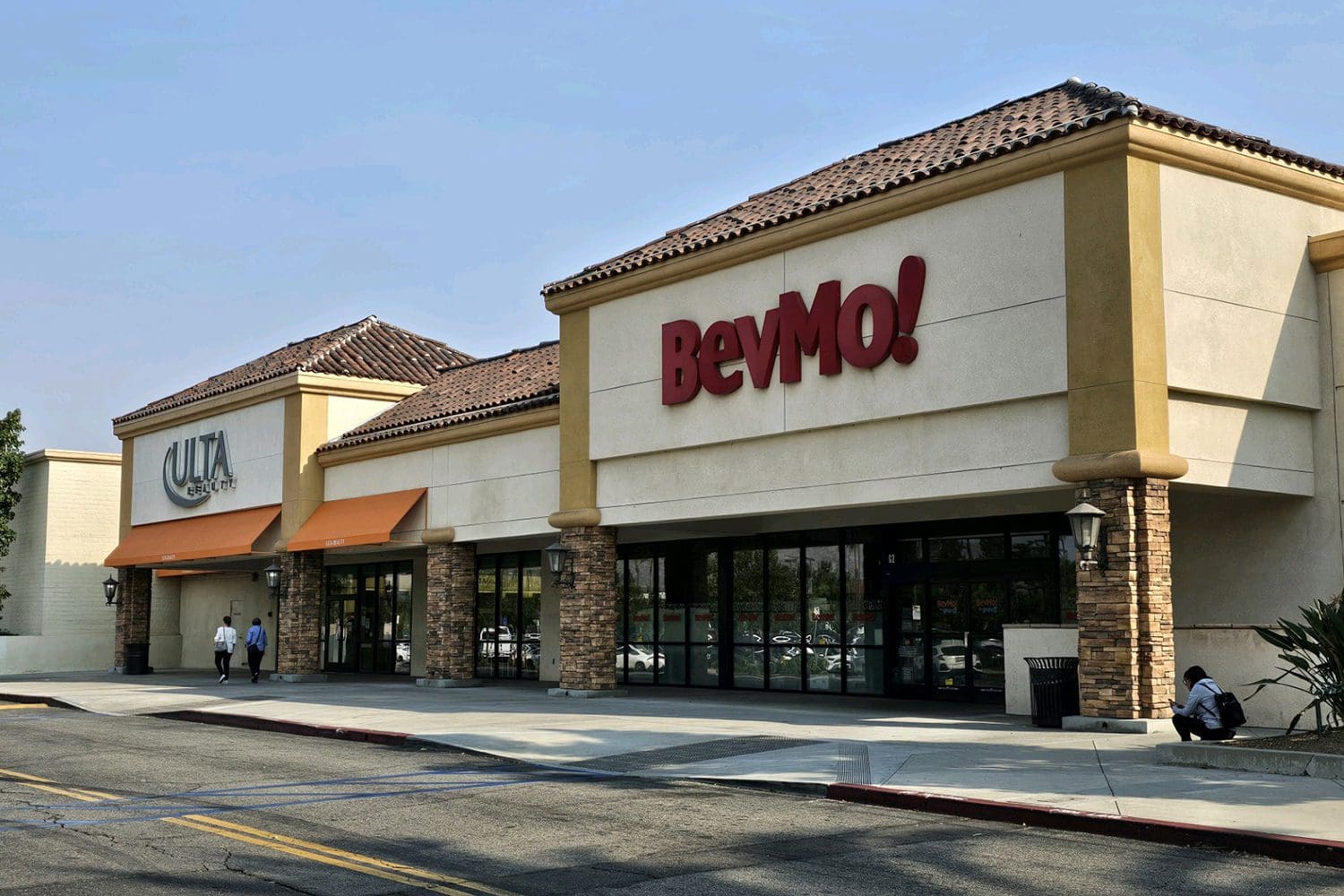 Storefronts of Ulta Beauty and BevMo! in a shopping center. A person is sitting near the entrance of BevMo! and two people are walking past Ulta Beauty.