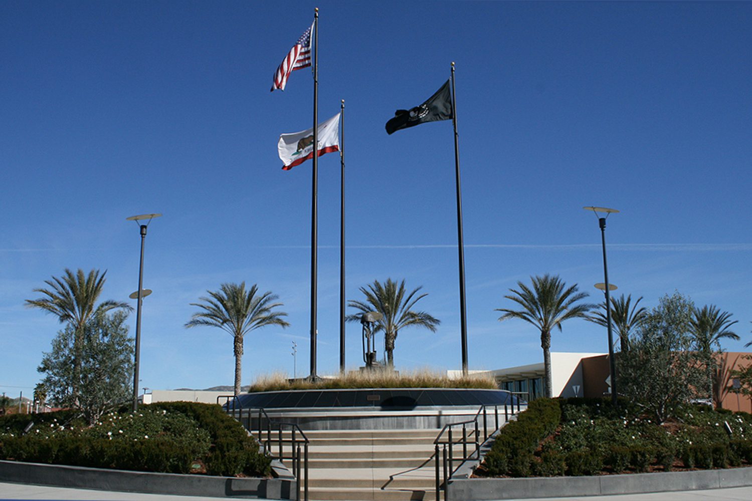 Three flagpoles with the US, California, and black POW/MIA flags stand in a plaza with palm trees and landscaped plants.