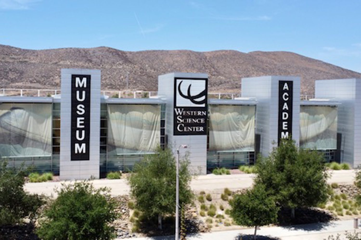 A large building labeled "Western Science Center" with banners reading "Museum" and "Academy." The building is surrounded by trees and desert hills in the background.