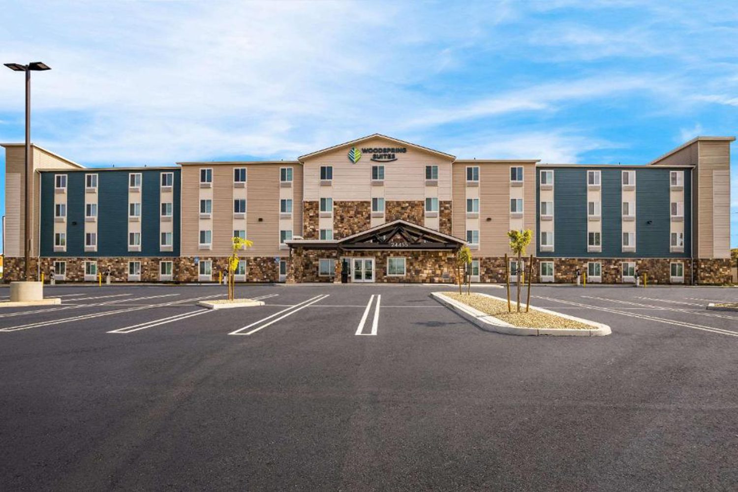 A four-story hotel building with a stone facade and green and gray panels, featuring the "WoodSpring Suites" sign, ample parking space, and a cloudy blue sky backdrop.