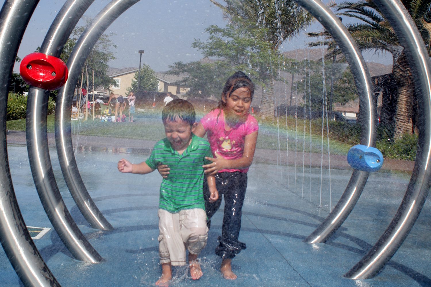 Two children playfully run through a water sprinkler feature at a park, with one child in a green shirt and the other in a pink shirt laughing and holding onto each other.