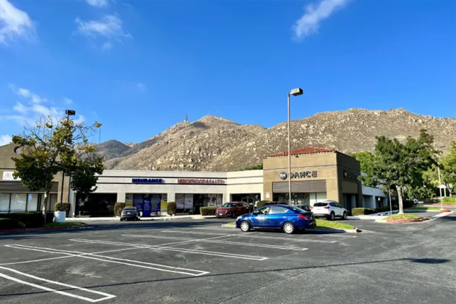 A parking lot in front of a strip mall with several shops, under a clear blue sky, and a mountain in the background. A few cars are parked in the lot.