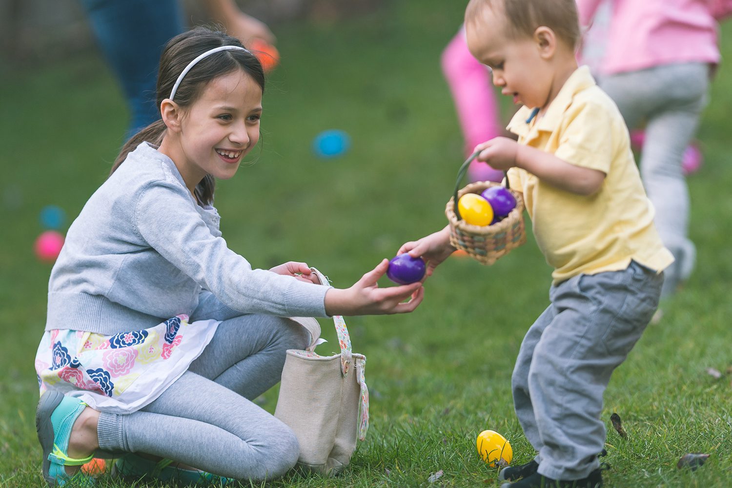 A young girl kneels on grass, smiling and handing a colorful egg to a toddler holding a basket, with more children in the background collecting eggs.