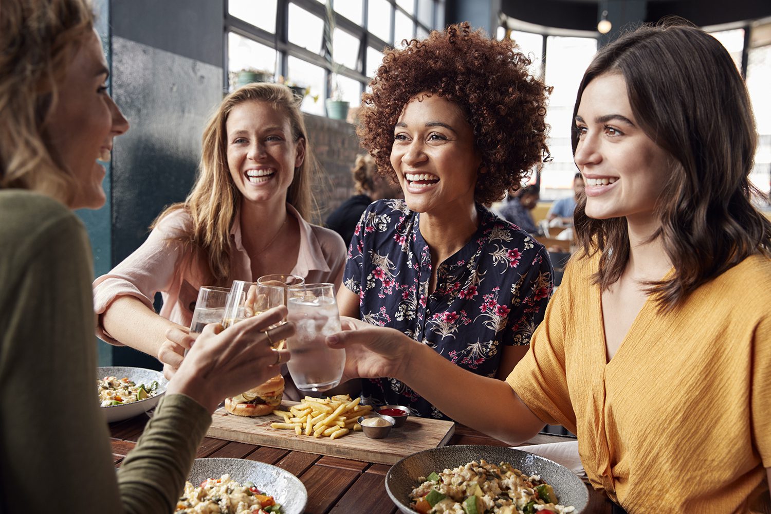 Four women sit at a table in a restaurant, smiling and holding up glasses in a toast. Plates of food, including salads and fries, are on the table in front of them.