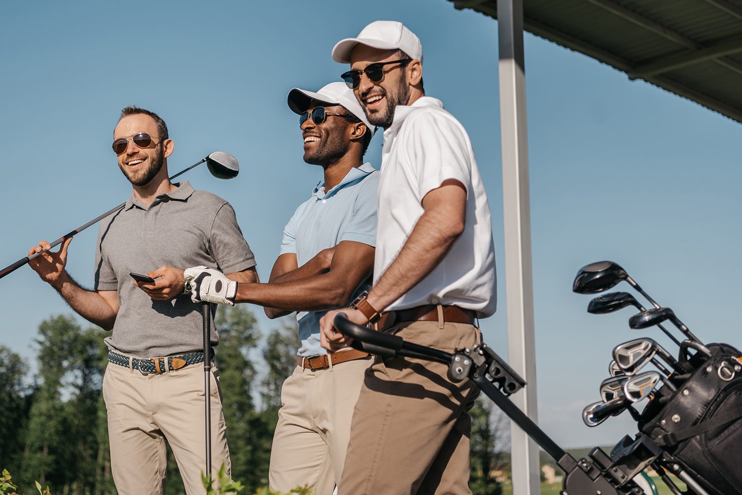 Three men wearing golf attire and sunglasses stand together outdoors at a golf course, smiling and holding golf equipment. Golf bags and clubs are visible beside them.