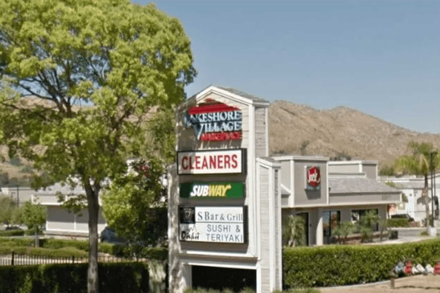 A signboard displays various business names including Lakeshore Village, Cleaners, Subway, S Bar & Grill, and a sushi and teriyaki restaurant. Buildings and a tree are visible in the background.