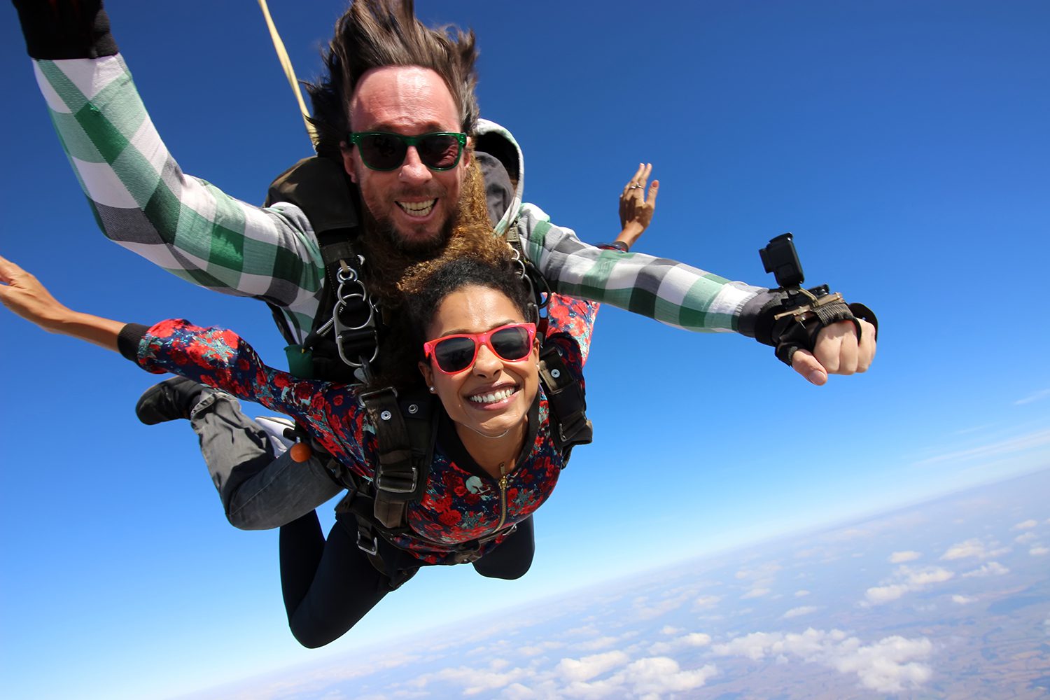 Two people are tandem skydiving, wearing harnesses and goggles. The instructor is behind, guiding the fall. The sky is clear and blue with a view of the clouds and landscape below.