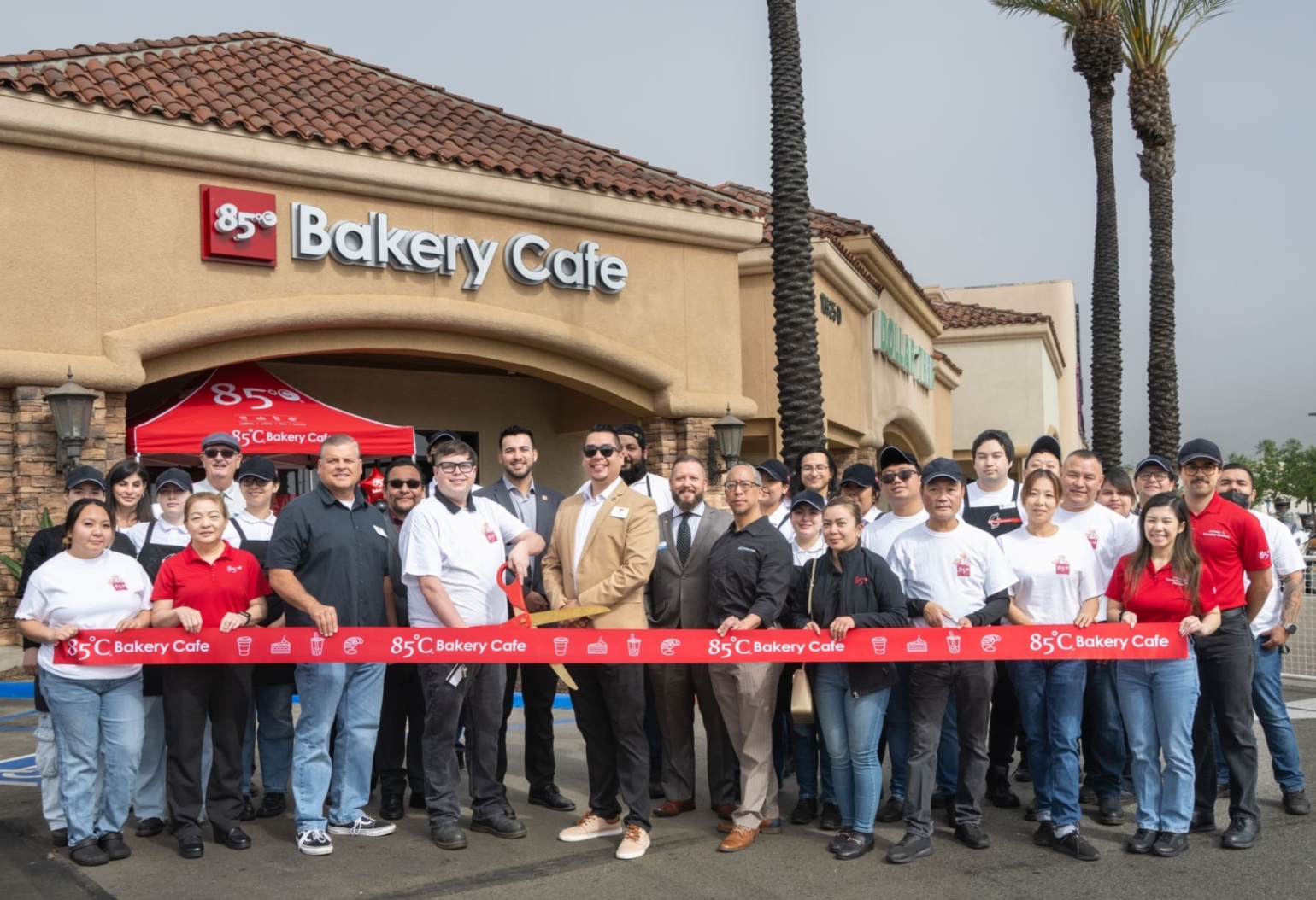 A group of people stand in front of a bakery cafe, holding a red ribbon during an outdoor event.