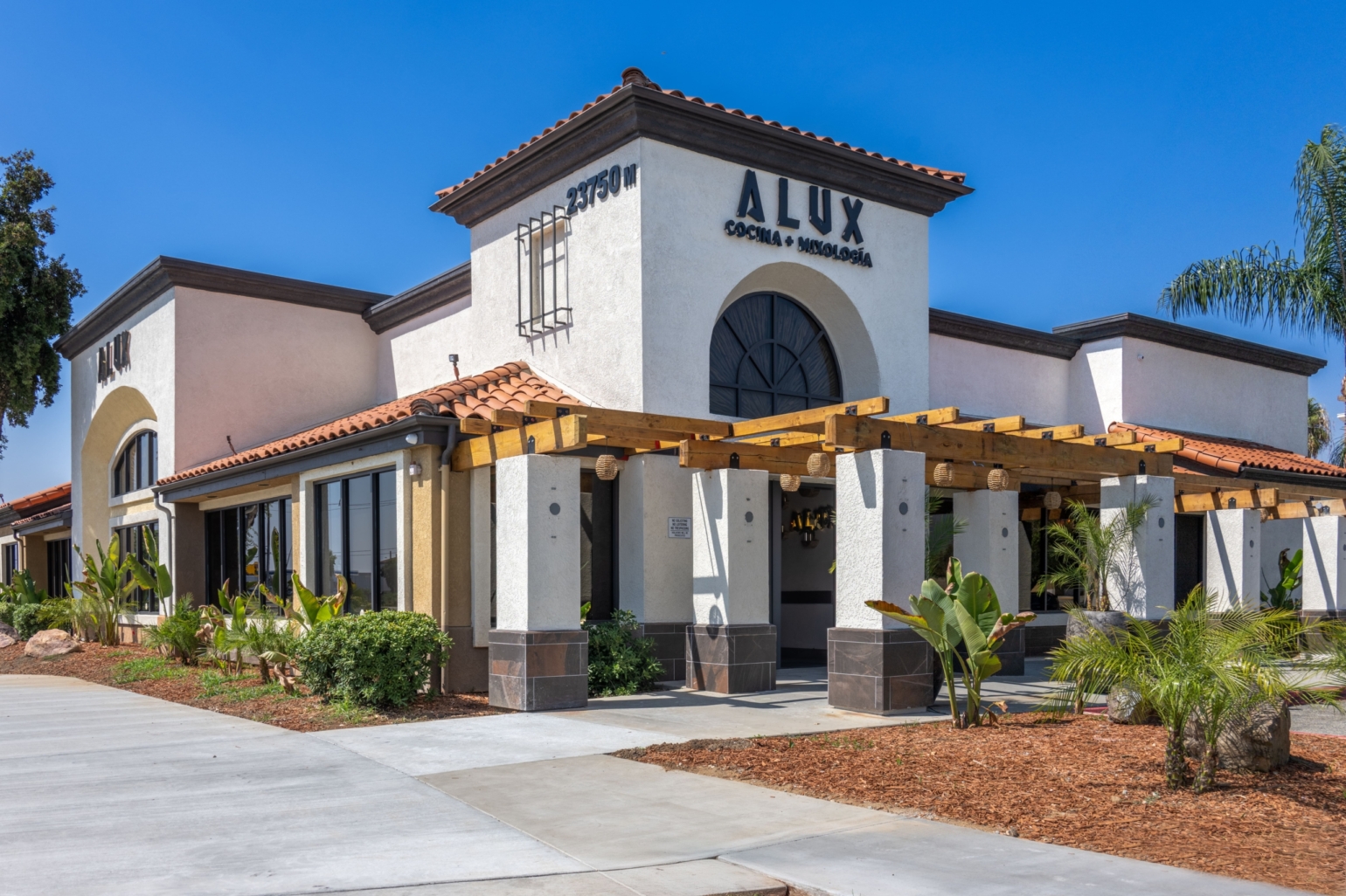 A modern restaurant with a Spanish-style exterior and pergola entrance, surrounded by palm trees and landscaping, on a sunny day.